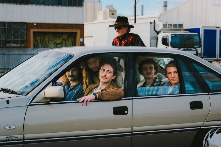 Band members of Outback Cadillac posing inside a vintage car, with a member wearing a cowboy hat standing behind it.